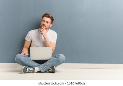 Young redhead student man sitting on the floor doubting and confused. He is holding a laptop.