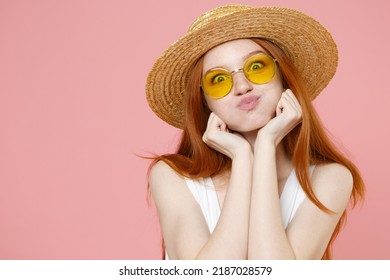 Young redhead woman ginger long hair in straw hat glasses summer clothes blow puff cheeks funny face mouth inflated air crazy expression prop up chin isolated on pastel pink background studio portrait
