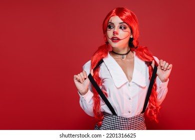 young redhead woman in halloween makeup touching suspenders and looking way isolated on red