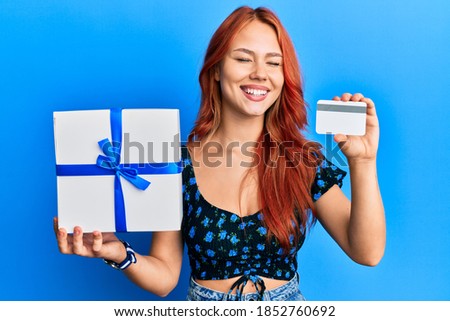Young redhead woman holding gift and credit card smiling and laughing hard out loud because funny crazy joke. 