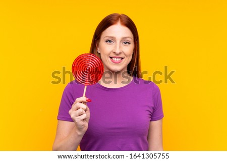 Young redhead woman holding a lollipop over yellow background smiling a lot