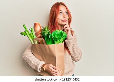 Young redhead woman holding paper bag with bread and groceries serious face thinking about question with hand on chin, thoughtful about confusing idea 
