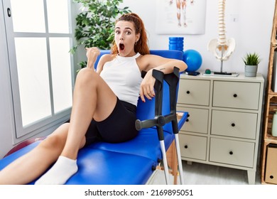 Young Redhead Woman Lying On Rehabilitation Bed Holding Crutches Surprised Pointing With Hand Finger To The Side, Open Mouth Amazed Expression. 