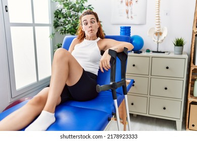 Young Redhead Woman Lying On Rehabilitation Bed Holding Crutches Making Fish Face With Lips, Crazy And Comical Gesture. Funny Expression. 