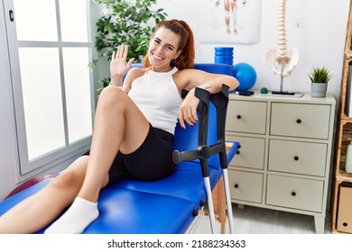 Young Redhead Woman Lying On Rehabilitation Bed Holding Crutches Waiving Saying Hello Happy And Smiling, Friendly Welcome Gesture 