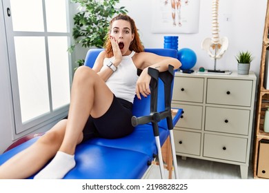 Young Redhead Woman Lying On Rehabilitation Bed Holding Crutches Afraid And Shocked, Surprise And Amazed Expression With Hands On Face 