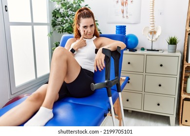 Young Redhead Woman Lying On Rehabilitation Bed Holding Crutches Pointing To The Eye Watching You Gesture, Suspicious Expression 