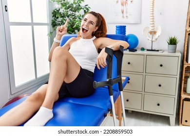 Young Redhead Woman Lying On Rehabilitation Bed Holding Crutches Smiling Amazed And Surprised And Pointing Up With Fingers And Raised Arms. 