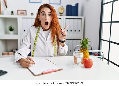 Young redhead woman nutritionist doctor at the clinic surprised pointing with finger to the side, open mouth amazed expression. 