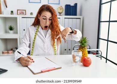 Young redhead woman nutritionist doctor at the clinic pointing down with fingers showing advertisement, surprised face and open mouth 