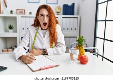 Young redhead woman nutritionist doctor at the clinic in shock face, looking skeptical and sarcastic, surprised with open mouth 