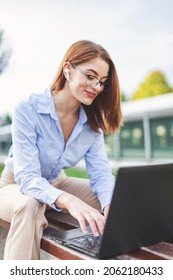 Young Redhead Woman Online Communicating In Park By Laptop And Hands Free Headset