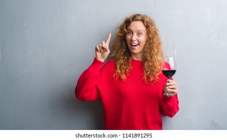 Young redhead woman over grey grunge wall drinking a glass of wine surprised with an idea or question pointing finger with happy face, number one - Powered by Shutterstock