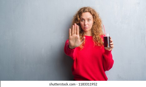 Young Redhead Woman Over Grey Grunge Wall Drinking Soda Refreshment With Open Hand Doing Stop Sign With Serious And Confident Expression, Defense Gesture