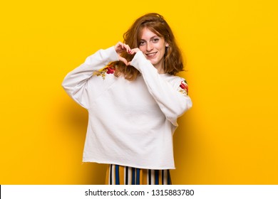 Young Redhead Woman Over Yellow Wall Making Heart Symbol By Hands