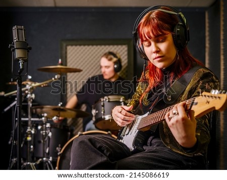 Young redhead woman playing electric guitar and a male drummer in a recording studio.