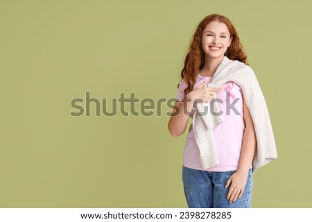Young redhead woman pointing at pink ribbon on green background. Breast cancer awareness concept