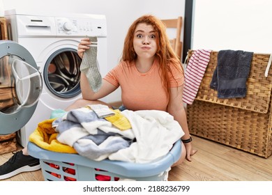 Young Redhead Woman Putting Dirty Laundry Into Washing Machine Puffing Cheeks With Funny Face. Mouth Inflated With Air, Crazy Expression. 