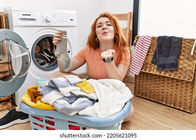 Young Redhead Woman Putting Dirty Laundry Into Washing Machine Looking At The Camera Blowing A Kiss With Hand On Air Being Lovely And Sexy. Love Expression. 