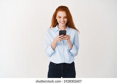 Young Redhead Woman Reading Message On Mobile Phone And Smiling. Businesswoman Looking Happy At Smartphone Screen, Standing Over White Background.