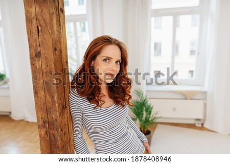 Young redhead woman scrutinising the camera with a knowing thoughtful expression as she leans against a wooden post indoors at home with copyspace