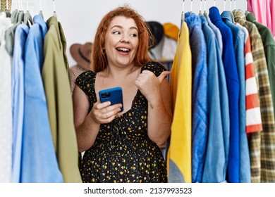 Young Redhead Woman Searching Clothes On Clothing Rack Using Smartphone Pointing Thumb Up To The Side Smiling Happy With Open Mouth 