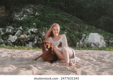 Young redhead woman sitting on a sandy beach near the ocean with a golden retriever and smiling. A woman gently strokes her dog and hugs it. Walking with your pet at dawn on a deserted beach. - Powered by Shutterstock