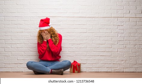 Young Redhead Woman Sitting Over Brick Wall Wearing Christmas Hat With Sad Expression Covering Face With Hands While Crying. Depression Concept.