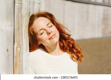 Young Redhead Woman Soaking Up The Sun On Her Face With A Blissful Smile And Closed Eyes As She Leans Against An Old Wall