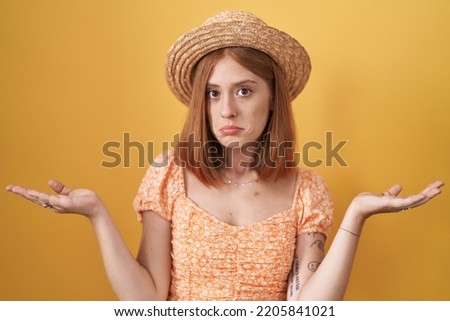 Young redhead woman standing over yellow background wearing summer hat clueless and confused expression with arms and hands raised. doubt concept. 