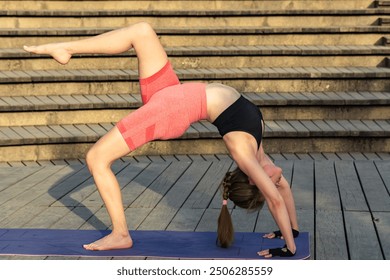 Young redhead woman stretching her body in a bridge pose during an outdoor yoga session. The scene has a relaxing atmosphere with steps in the background, on a wooden surface - Powered by Shutterstock