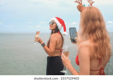 Young redhead woman taking photo of her brunette friend on her phone wearing Santa hat with glass of champagne in her hand against the ocean on vacation. Christmas in summer in tropics with friends. - Powered by Shutterstock