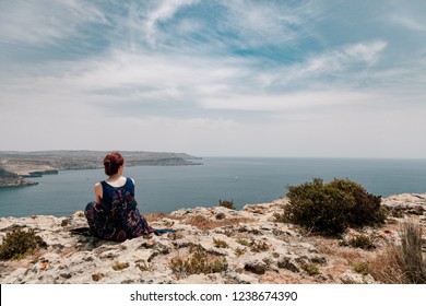 Young redhead woman with a topknot sitting on the edge of a cliff looking far away the sea