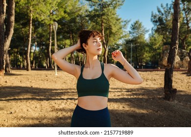 Young redhead woman touching hair and looking away, wearing green sports bra and blue yoga pants while standing on city park, outdoors. Healthy life and outdoor sports concept.