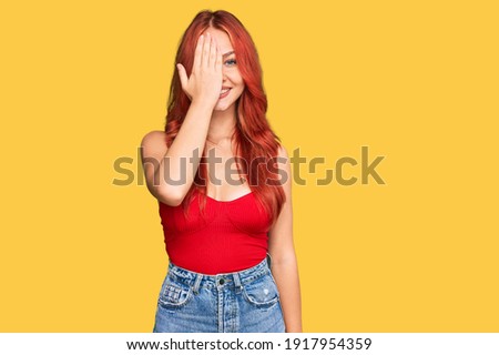 Young redhead woman wearing casual clothes covering one eye with hand, confident smile on face and surprise emotion. 
