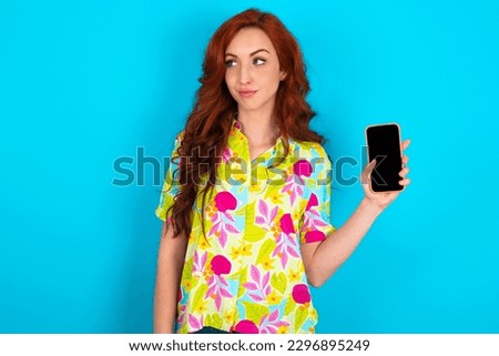 Young redhead woman wearing colorful shirt over blue background holds new mobile phone and looks mysterious aside shows blank display of modern cellular