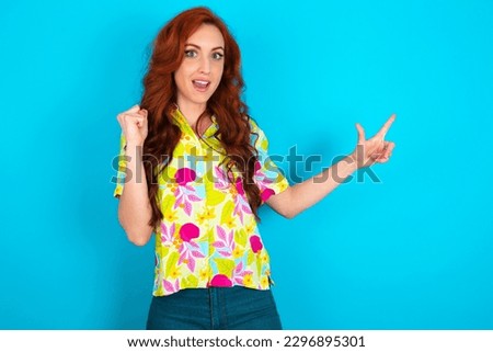 Young redhead woman wearing colorful shirt over blue background points at empty space holding fist up, winner gesture.