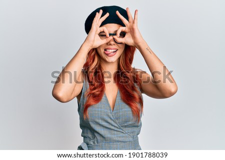 Young redhead woman wearing fashion french look with beret doing ok gesture like binoculars sticking tongue out, eyes looking through fingers. crazy expression. 