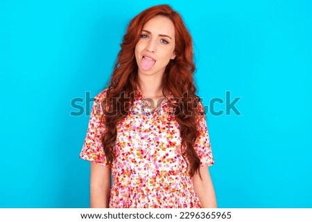 Young redhead woman wearing floral dress over blue background with happy and funny face smiling and showing tongue.