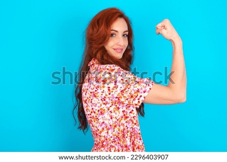 Young redhead woman wearing floral dress over blue background ,  showing muscles after workout. Health and strength concept.
