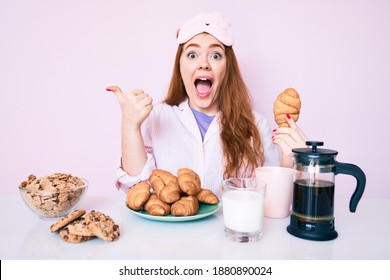 Young Redhead Woman Wearing Sleep Mask And Pajama Eating Breakfast Holding Croissant Pointing Thumb Up To The Side Smiling Happy With Open Mouth 