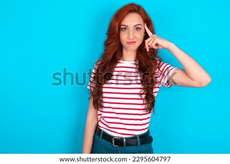 young redhead woman wearing striped T-shirt over blue background tries to memorize something, keeps fore finger on temple, reminds information for exam,