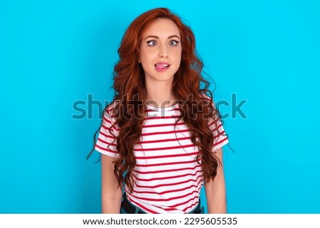 young redhead woman wearing striped T-shirt over blue background showing grimace face crossing eyes and showing tongue. Being funny and crazy