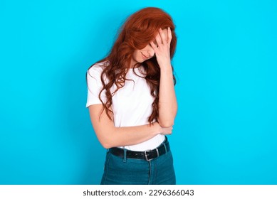 Young redhead woman wearing white T-shirt over blue background making facepalm gesture while smiling amazed with stupid situation. - Powered by Shutterstock