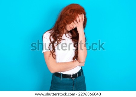 Young redhead woman wearing white T-shirt over blue background making facepalm gesture while smiling amazed with stupid situation.