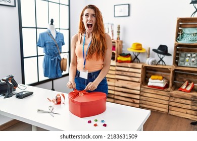 Young redhead woman working as manager at retail boutique wrapping gift angry and mad screaming frustrated and furious, shouting with anger looking up. 