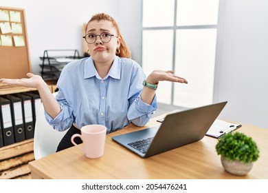 Young Redhead Woman Working At The Office Using Computer Laptop Clueless And Confused Expression With Arms And Hands Raised. Doubt Concept. 