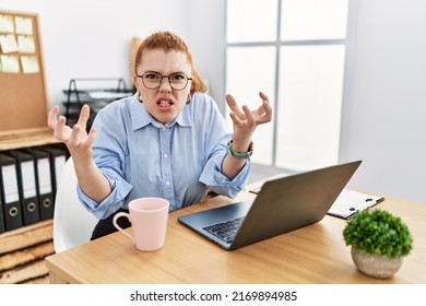 Young redhead woman working at the office using computer laptop shouting frustrated with rage, hands trying to strangle, yelling mad 