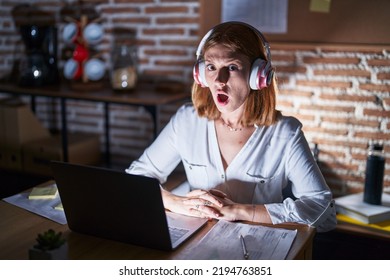 Young Redhead Woman Working At The Office At Night Wearing Headphones In Shock Face, Looking Skeptical And Sarcastic, Surprised With Open Mouth 