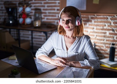 Young Redhead Woman Working At The Office At Night Wearing Headphones Smiling Looking To The Side And Staring Away Thinking. 